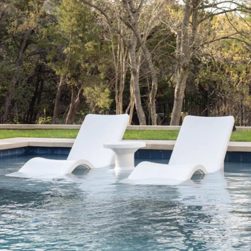 Two white tanning ledge chairs and a side table partially submerged in a pool, surrounded by greenery.