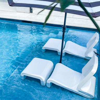 Two white tanning ledge chairs and matching ottomans partially submerged in the pool ledge under a large striped umbrella.