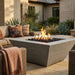 A modern square concrete fire pit table with black river rocks and active orange flames, set on a stone patio in an outdoor patio lounge area with wicker furniture and cushions.