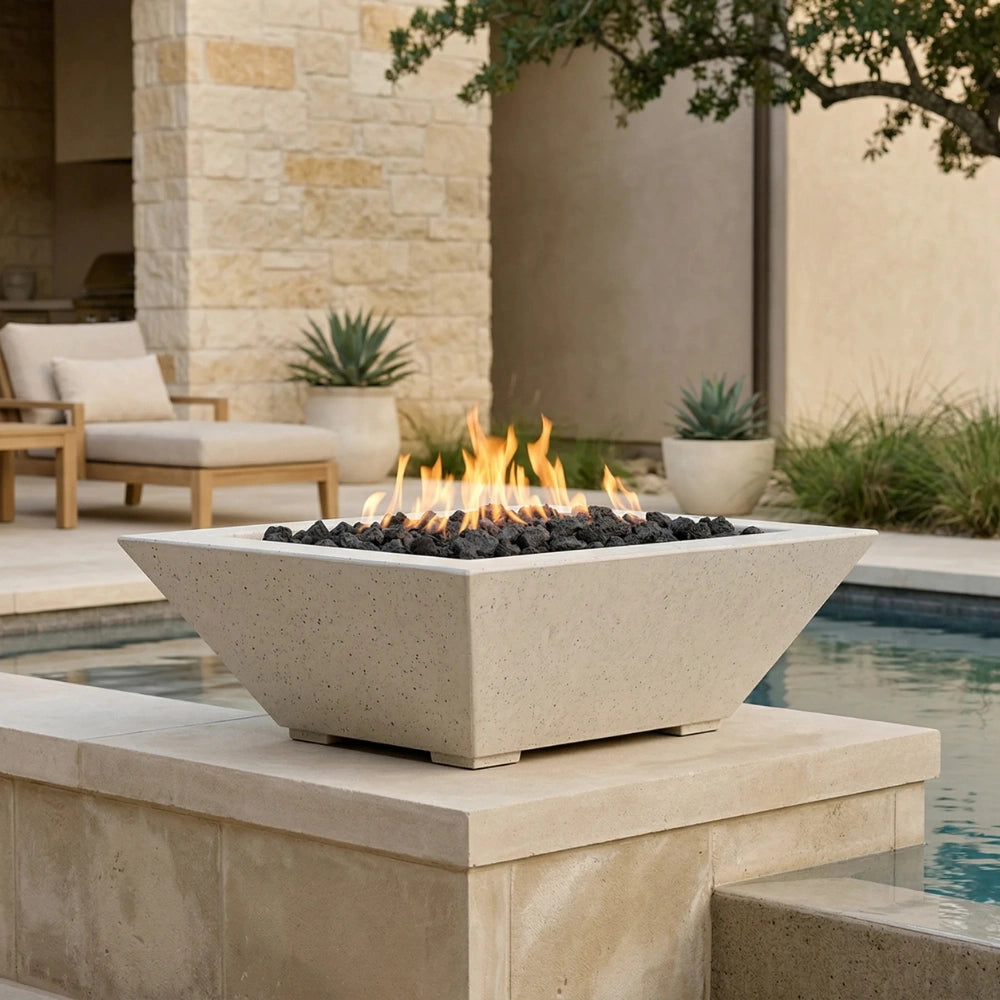 A modern square concrete fire pit with black river rocks and active orange flames, set on a stone patio next to a swimming pool.