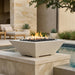 A modern square concrete fire pit with black river rocks and active orange flames, set on a stone patio next to a swimming pool.