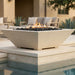 A modern square concrete fire pit table with black river rocks and active flames, set on a stone pool deck in an outdoor patio environment.