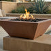 A square concrete fire pit with a warm terracotta-brown finish, filled with black lava rocks and active orange flames, set on a stone pedestal in an outdoor patio environment.