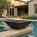 A modern black square fire pit table with a geometric slanted design, filled with dark river rocks and active flames, situated on a stone platform next to a swimming pool in a luxurious backyard setting.