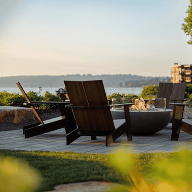 Four black outdoor lounge chairs with wood grain backrest placed around a concrete round outdoor fire pit bowl on an outdoor patio setting