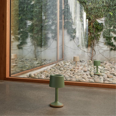 small green outdoor lamp on the floor in front of a glass wall, with pebbles outside d two similar lamps in green and beige