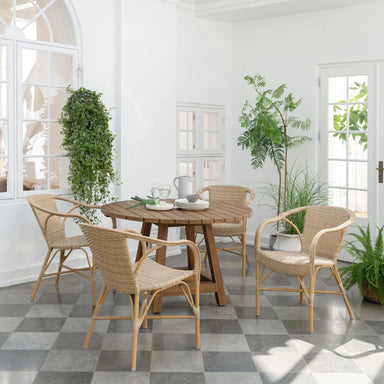 Natural rattan outdoor dining armchair beside a matching round wooden dining table in a bright indoor setting.