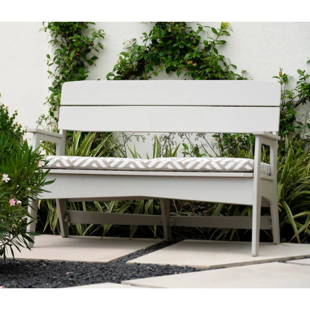 A white outdoor bench with a cushioned seat, set against a white wall with climbing green plants and garden foliage.