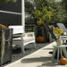 Outdoor patio scene with a white wooden bench, dark lounge chairs, potted plants, pumpkins, and a small mint-green side table on a concrete walkway.