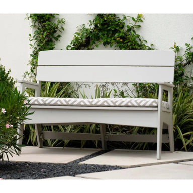 A white outdoor bench with a cushioned seat, set against a white wall with climbing green plants and garden foliage.