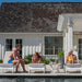 Three people relax on white lounge chairs by a swimming pool in front of a white house with a dark shingled roof. The scene is bright and sunny, with the pool deck, plants, and patio furniture visible.