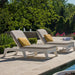 Two outdoor chaise lounge chairs with light beige cushions and yellow accent pillows, positioned on a stone patio poolside with lush green trees in the background.