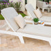 Two white outdoor lounge chaise lounges with light beige cushions and patterned throw pillows on a stone patio, with a small side table holding a plant and books between them.