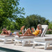 Two women relax on white outdoor chaise lounge chairs beside a swimming pool on a sunny day, with trees and blue sky in the background.