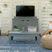 A gray outdoor TV stand with a mounted flat-screen television, set in a patio lifestyle scene with a matching gray coffee table, blue patterned rug, and a person sitting on a woven pouf in the background.