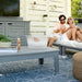 A man and woman relax on a white outdoor chaise lounge on a patio, with a large clear ice bucket in the foreground. The setting includes a gray coffee table, blue patterned rug, potted plants, and a white slatted wall backdrop.