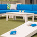 Outdoor patio furniture set featuring a bright blue sectional sofa, a white rectangular coffee table, and a matching white foreground table with blue and white bean bags, set on green artificial turf.