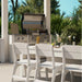 Outdoor patio scene with a stainless steel gas grill mounted above a matching grill cabinet, set beside a white dining table and chairs with green plants and trees in the background.