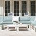 An outdoor patio seating set featuring a light aqua blue sectional sofa with white frames, a matching white coffee table, and white patio chairs in front of white French doors.