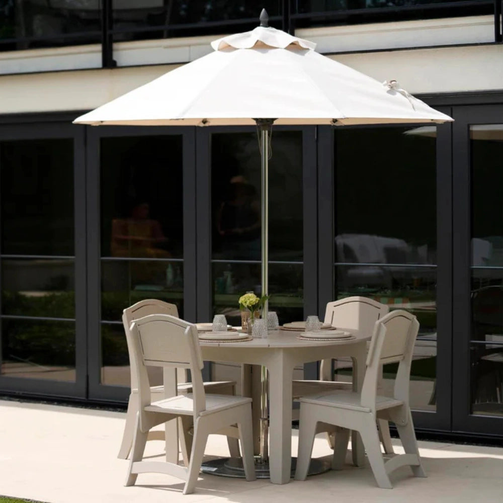 An outdoor patio dining set featuring a round light beige table, four matching armchairs, and a large cream patio umbrella with a silver pole, set against a modern glass door background.