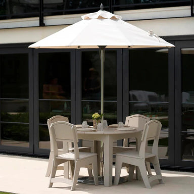 An outdoor patio dining set featuring a round light beige table, four matching armchairs, and a large cream patio umbrella with a silver pole, set against a modern glass door background.