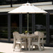 An outdoor patio dining set featuring a round light beige table, four matching armchairs, and a large cream patio umbrella with a silver pole, set against a modern glass door background.