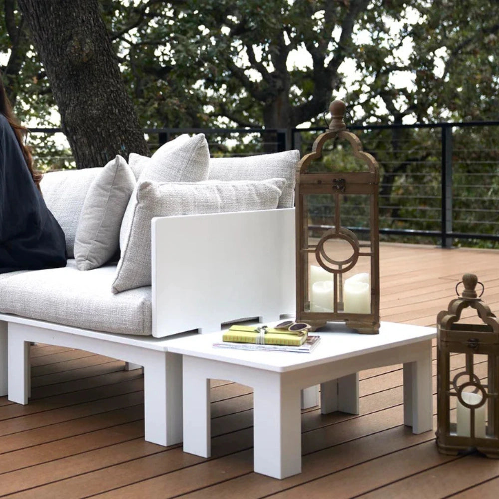 White outdoor patio furniture set on a wooden deck, featuring a lounge chair with light gray cushions, a matching low coffee table, and two decorative lanterns against a backdrop of trees.