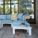A light blue sectional outdoor sofa with a white frame on a patio, styled with a yellow and white patterned accent pillow, a white coffee table, and a small plant on a tray.