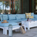 Outdoor sectional sofa set with light blue textured cushions and white frames, styled on a patio with a white coffee table, patterned pillows, a straw hat, and a woven bag.