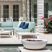 A light blue outdoor sectional sofa with navy and white pillows, paired with white outdoor tables and a white fire pit in a patio setting in front of a white house.