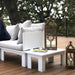 White outdoor patio furniture set on a wooden deck, featuring a lounge chair with light gray cushions, a matching low coffee table, and two decorative lanterns against a backdrop of trees.