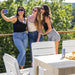 Three women taking a selfie on a sunny outdoor patio with white chairs and a resin table in the foreground, with lush green trees in the background.