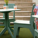 A mint green outdoor dining set consisting of a square table and two matching chairs, arranged on green artificial turf in front of beige stone steps. A small glass vase with yellow flowers and two patterned pink drinks sit on the table.