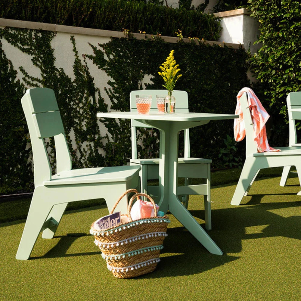 A mint green outdoor patio dining set consisting of a square table and two matching chairs on artificial grass, styled with a pink towel, a yellow flower vase, and a woven basket in the foreground.