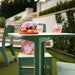 A mint green outdoor patio dining set with a square table and matching bench, styled in a garden setting with a striped blanket, fruit, and textured drinking glasses.