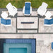 Overhead view of an outdoor patio setup with six white Adirondack-style chairs, four with blue cushions, arranged around a rectangular fire pit on light stone pavers. A swimming pool and grass are visible nearby.