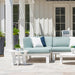 A light blue outdoor sectional sofa with white and navy throw pillows, set on a stone patio in front of a white house with French doors. A matching white side table sits in the foreground.