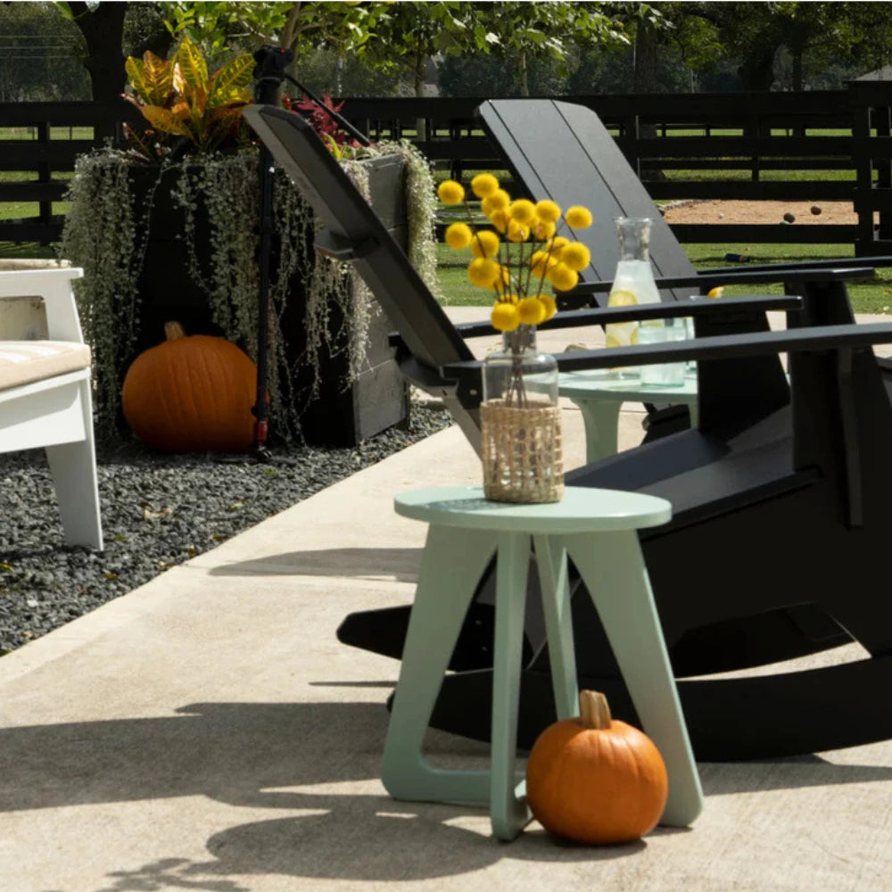 A mint green plastic side table in an outdoor patio setting, styled with a glass vase of yellow flowers and a small orange pumpkin on its base, next to black lounge chairs.