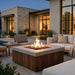 A modern outdoor fire pit table with a square concrete top and wood-look base, lit with flames in a patio setting. Comfortable beige lounge chairs, a side table, and a contemporary house with warm windows are visible in the background at dusk.