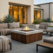 A modern outdoor patio scene centered on a square fire pit table with a concrete top and dark wood slat sides, surrounded by gray upholstered seating and plants in front of a contemporary house.