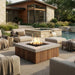 A modern square outdoor fire pit table with a wood-look slatted base and a light stone top, shown lit in a patio setting with lounge chairs, ottomans, a pool, and a house in the background.