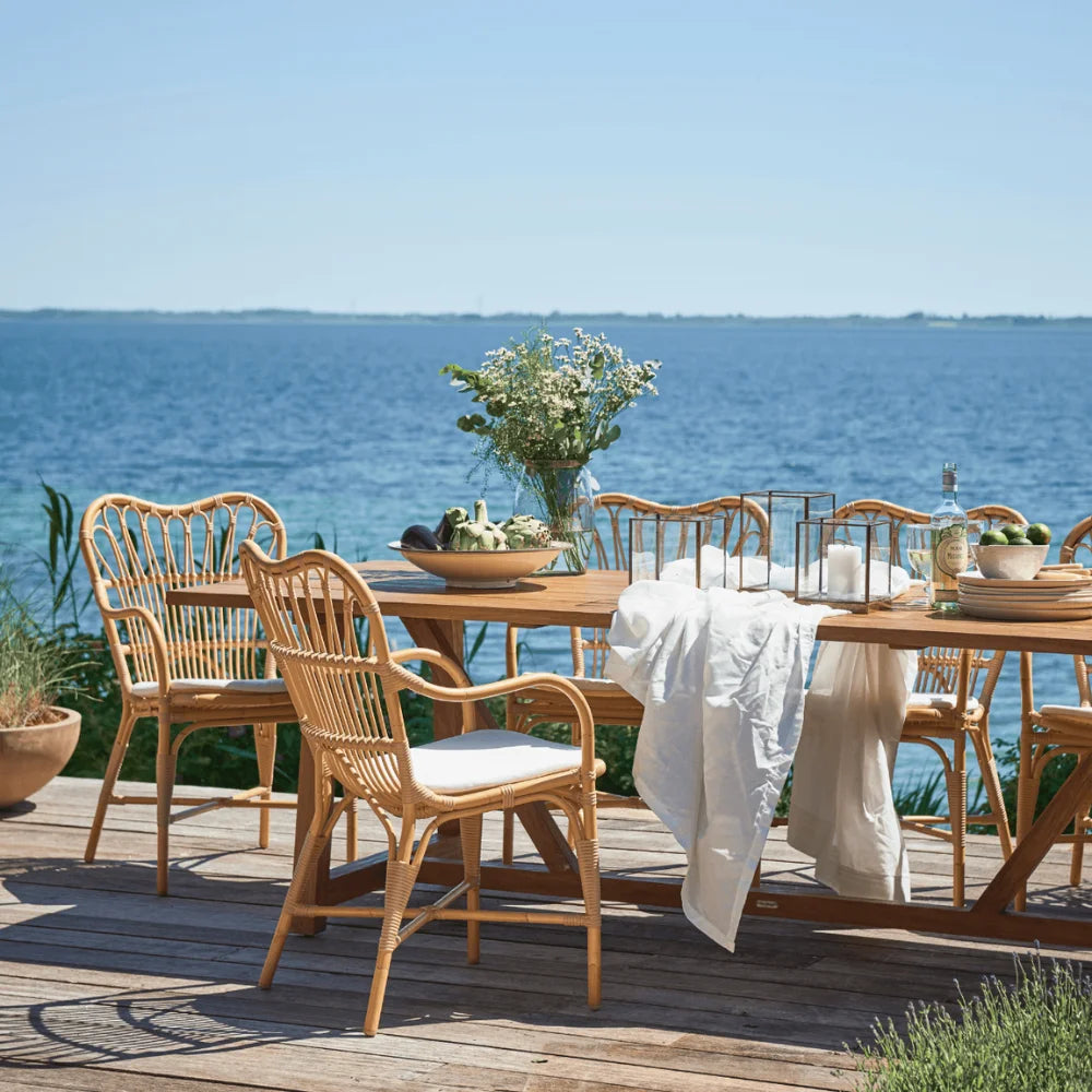 Natural rattan outdoor dining armchair with white cushions beside a matching wooden dining table by the ocean.