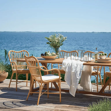 Natural rattan outdoor dining armchair with white cushions beside a matching wooden dining table by the ocean.