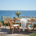 Natural rattan outdoor dining armchair with white cushions beside a matching wooden dining table by the ocean.