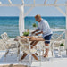 White rattan outdoor dining armchair beside a matching wooden dining table with a person on a seaside patio.