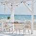 White rattan outdoor dining armchairs beside a matching square bistro table on a seaside patio.