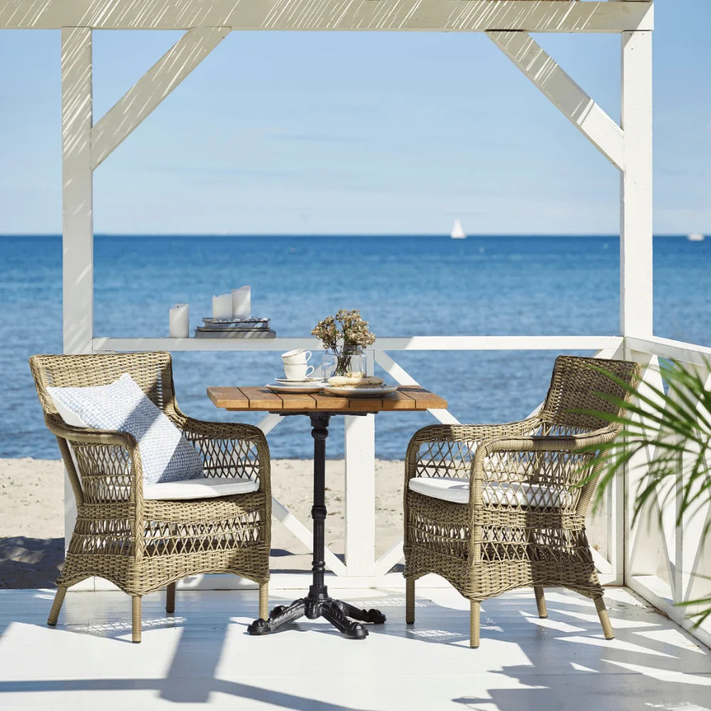 Gray rattan outdoor dining armchairs with white cushions beside a matching square bistro table by the ocean.