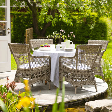 Gray rattan outdoor dining armchairs with white cushions around a matching round dining table in a garden.