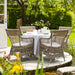 Gray rattan outdoor dining armchairs with white cushions around a matching round dining table in a garden.