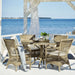 Gray rattan outdoor dining armchairs with white cushions around a matching round dining table by the ocean.